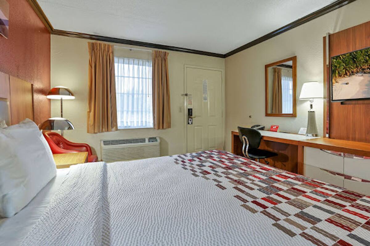 A hotel room with a bed, a desk, a chair, a desk lamp, a telephone, a mirror, and a window with curtains.