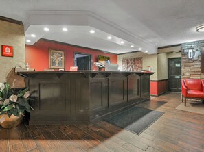 A reception area with a dark wooden reception desk, a red wall with artwork, a door, and a red leather chair. - Red Roof Inn Uhrichsville (Uhrichsville)