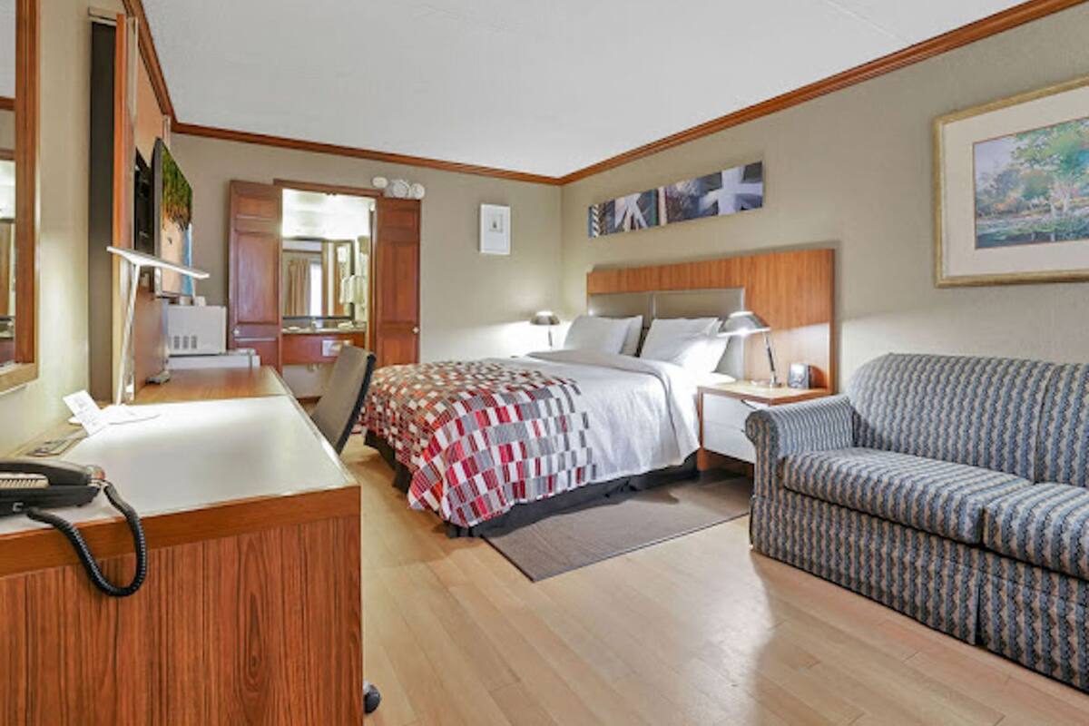 A hotel room with a bed, a desk with a telephone, a chair, a sofa, and a painting on the wall.
