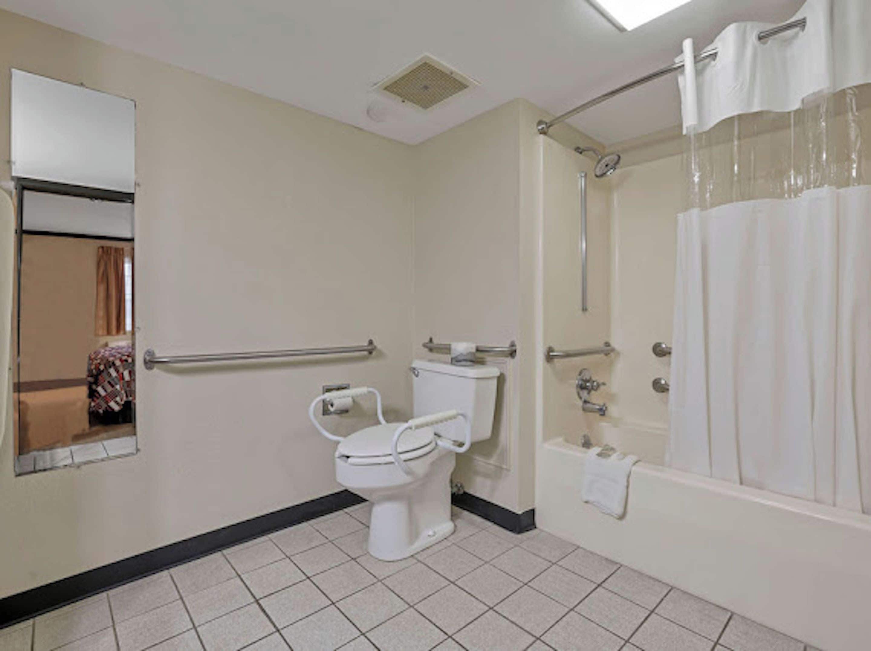 A bathroom with a toilet, a shower with a curtain, a handrail, and a mirror.