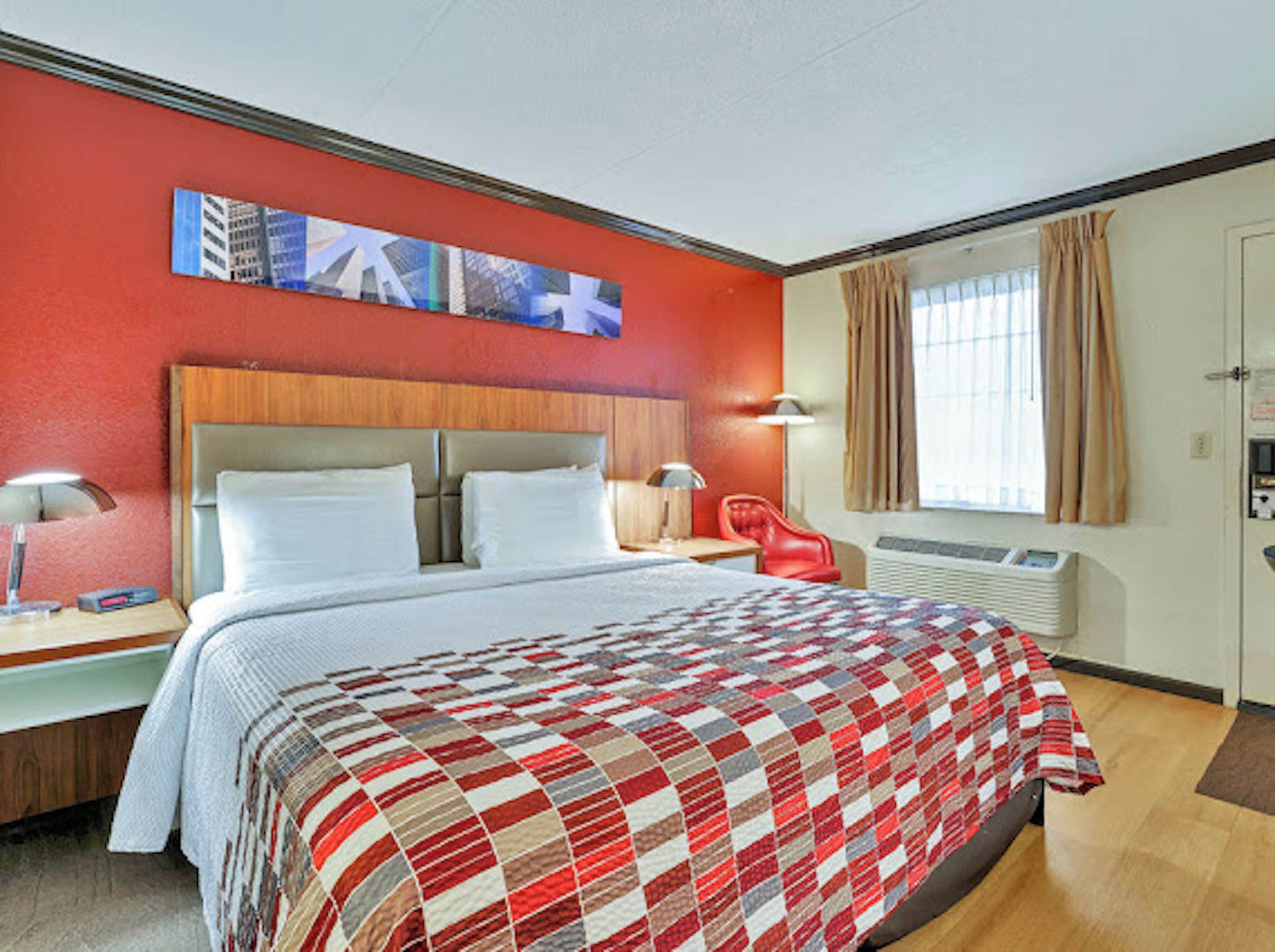 A hotel room with a bed, a red chair, a desk with a lamp, a wall-mounted air conditioner, and a window with curtains.