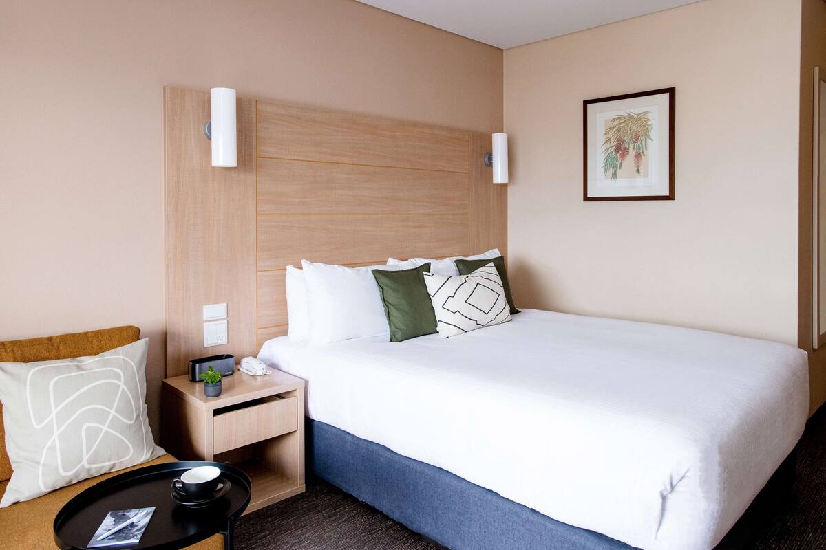A hotel room with a large bed, a bedside table with a lamp, a small plant, and a framed picture on the wall.