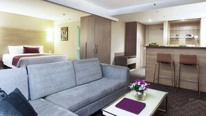 Habitación de hotel con un sofá gris, una pequeña mesa con un florero, una cama con almohadas, una kitchenette con barra y un baño visible al fondo.