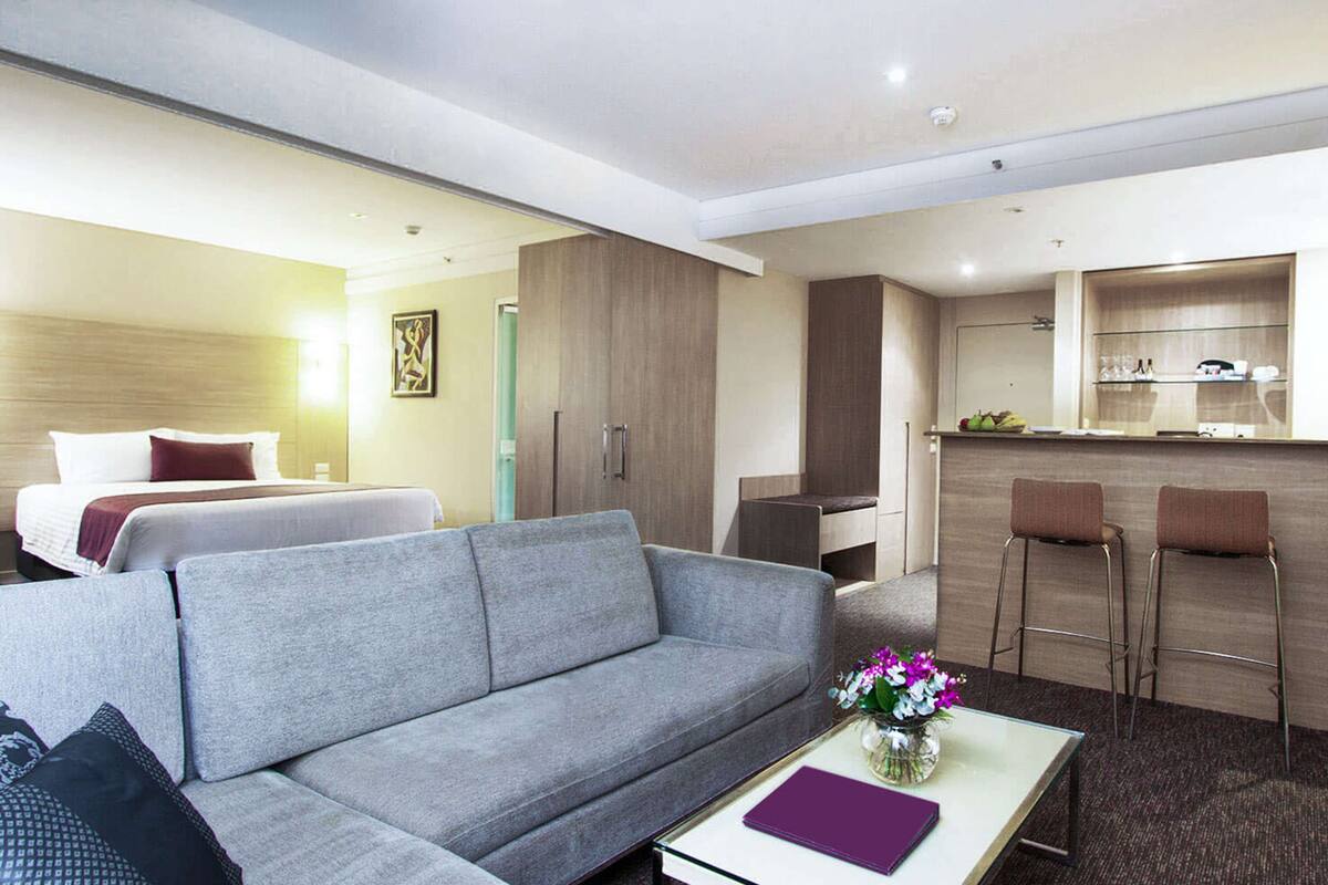 A hotel room with a grey sofa, a small table with a vase of flowers, a bed with pillows, a kitchenette with a bar area, and a bathroom visible in the background.