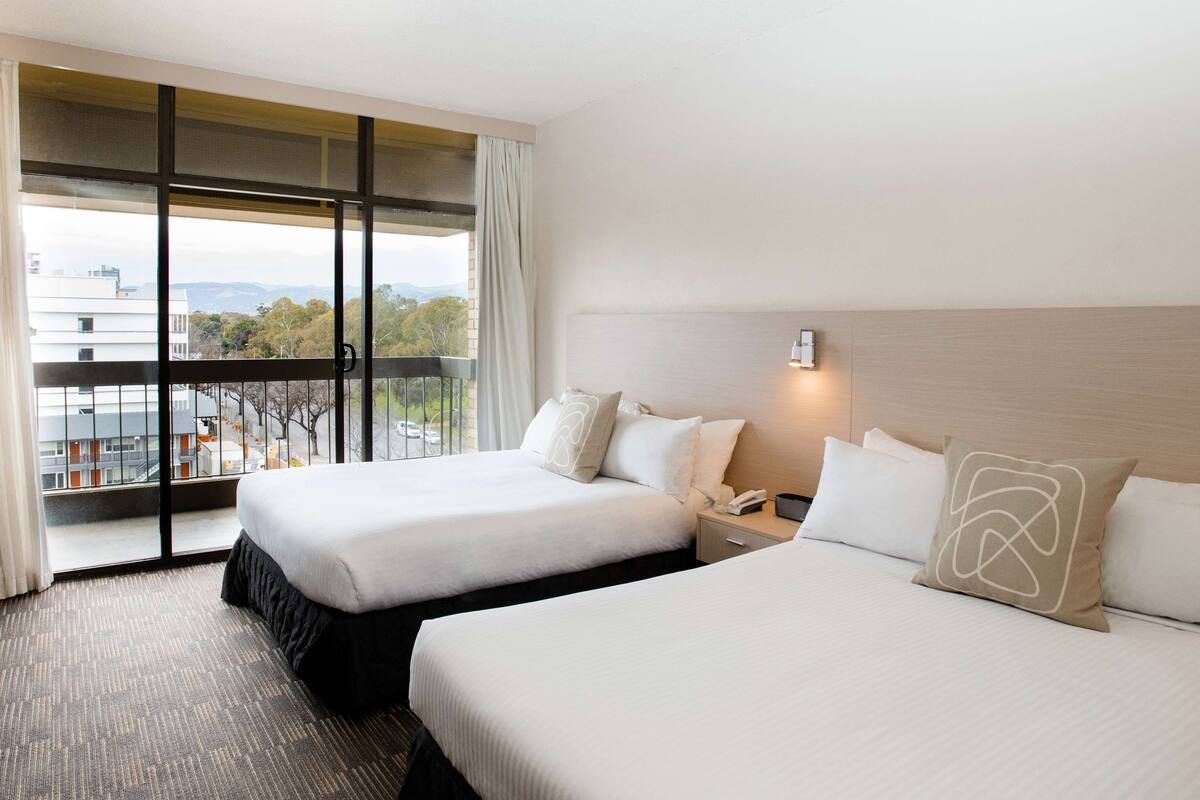 A hotel room with two beds, a large window with a view of buildings and trees, and a nightstand with a phone and lamp.