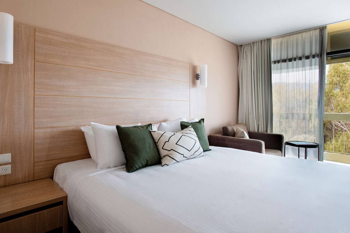 A hotel room with a large bed, a nightstand, a chair, a small table, and a window with curtains.