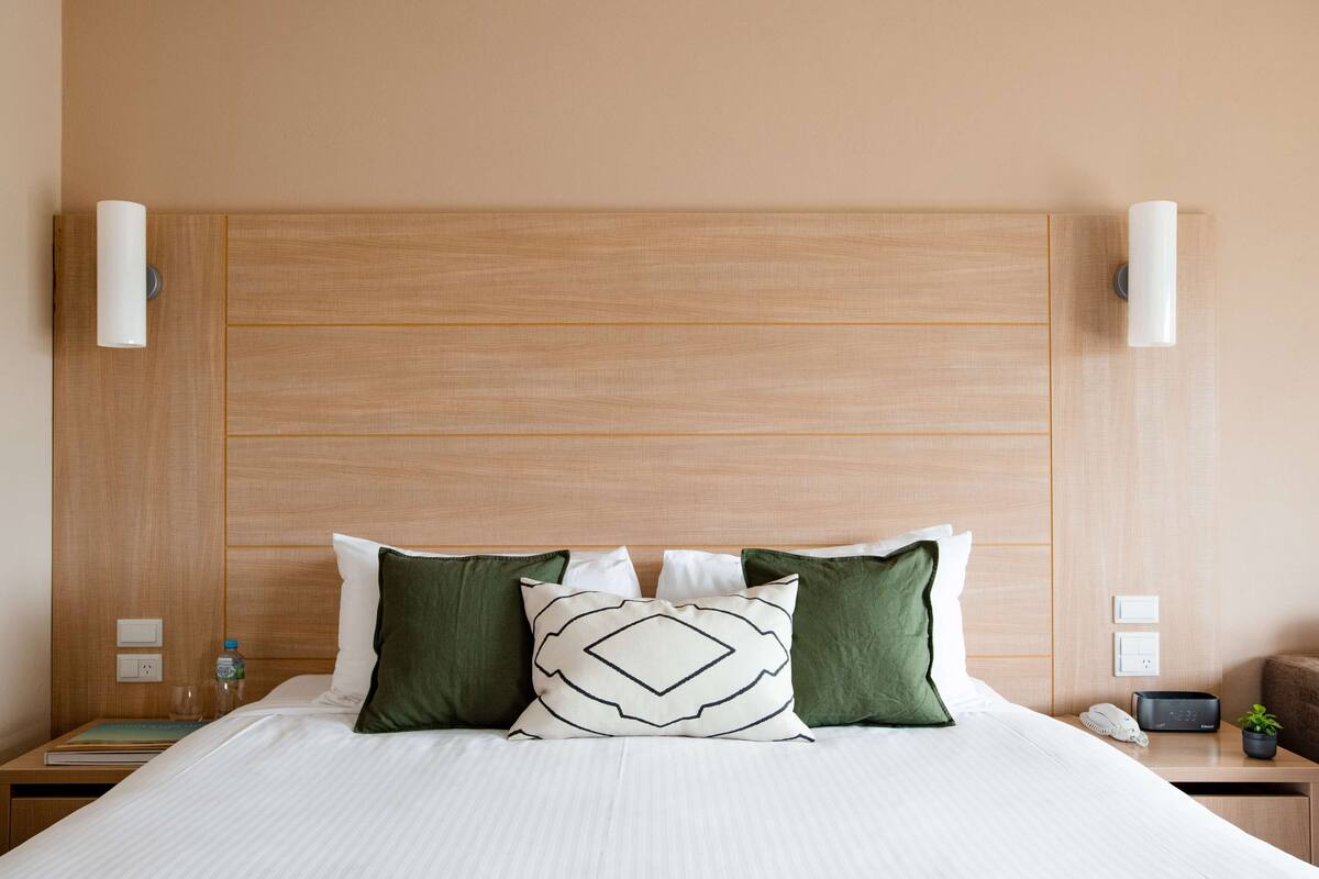 A hotel room with a wooden headboard, a bed with white linens, green and white pillows, a bedside table with a lamp, a phone, and a small plant.