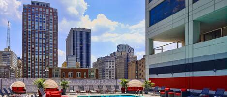 A rooftop pool with lounge chairs, a view of the city skyline, and a modern building with large windows.