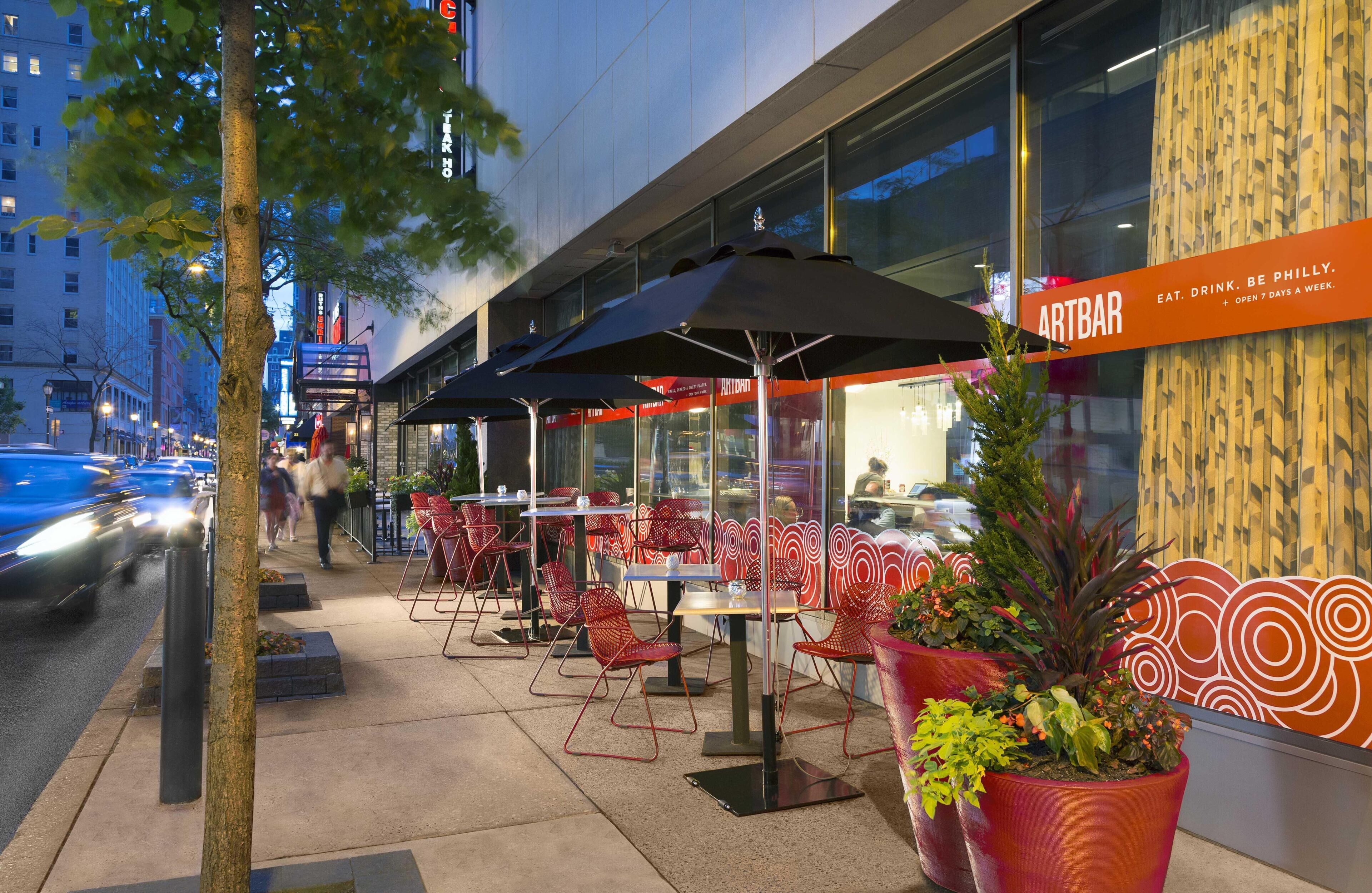 An outdoor seating area with tables and chairs, a sign for Artbar, and a tree in the foreground.