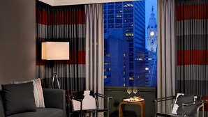 Een moderne hotelkamer met een bank, twee fauteuils, een bijzettafeltje met een vaas, een lamp en uitzicht op een stadsgezicht met een klokkentoren.