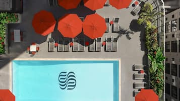 Eine Vogelperspektive auf ein Hotelpool-Areal mit mehreren roten Sonnenschirmen, Liegestühlen und einem großen Pool mit einem Logo.