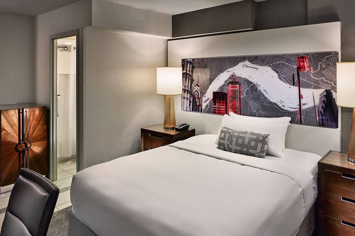 Une chambre d’hôtel avec un grand lit, une table de chevet avec une lampe, une chaise et une vue sur le paysage urbain par la fenêtre.