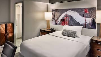 A hotel room with a large bed, a nightstand with a lamp, a chair, and a view of a cityscape through the window.