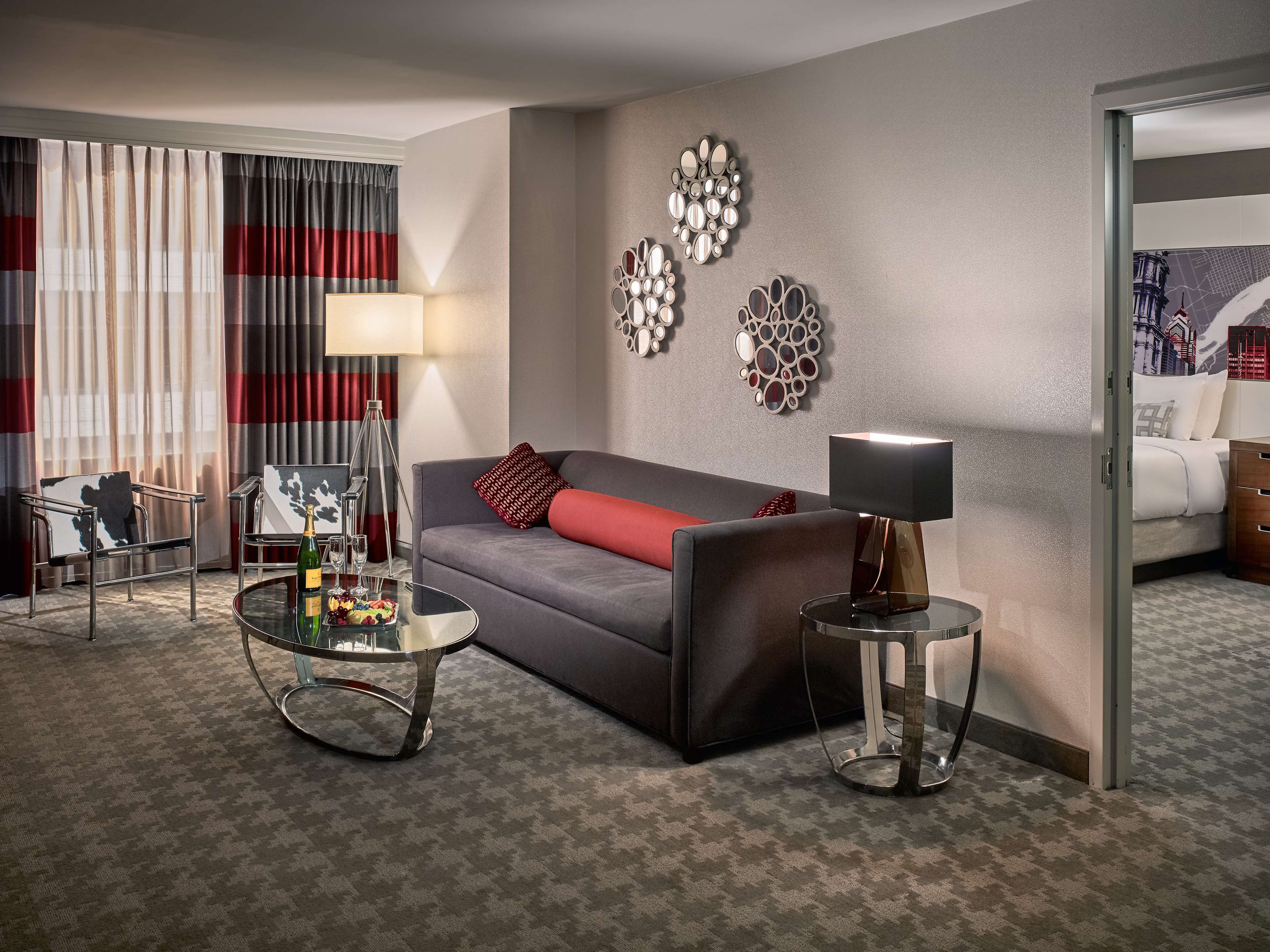 Une chambre d’hôtel avec un canapé gris, une table basse en verre, une table d’appoint avec une lampe, un miroir au mur et un lit recouvert d’une couette blanche.