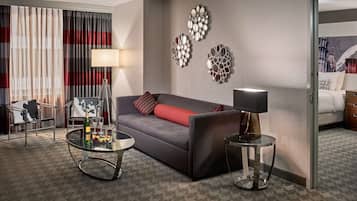 Een hotelkamer met een grijze bank, een glazen salontafel, een nachtkastje met een lamp, een spiegel aan de muur en een bed met een witte dekbedovertrek.
