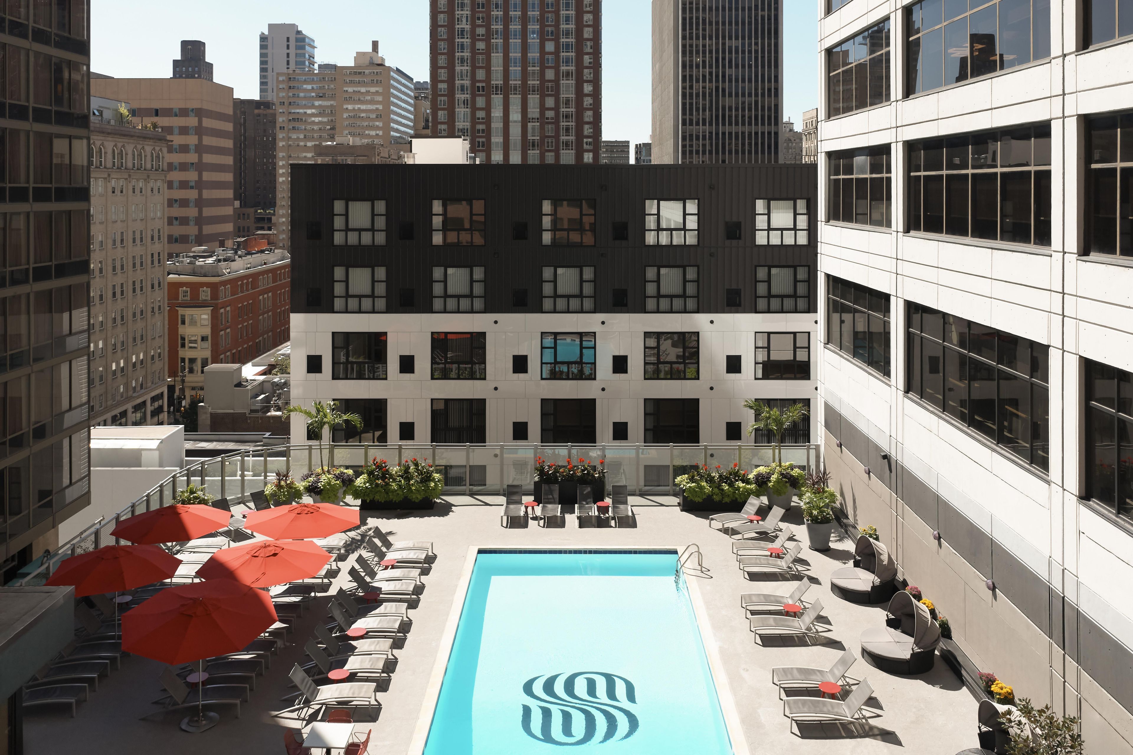 A rooftop pool area with lounge chairs and umbrellas, surrounded by modern buildings.