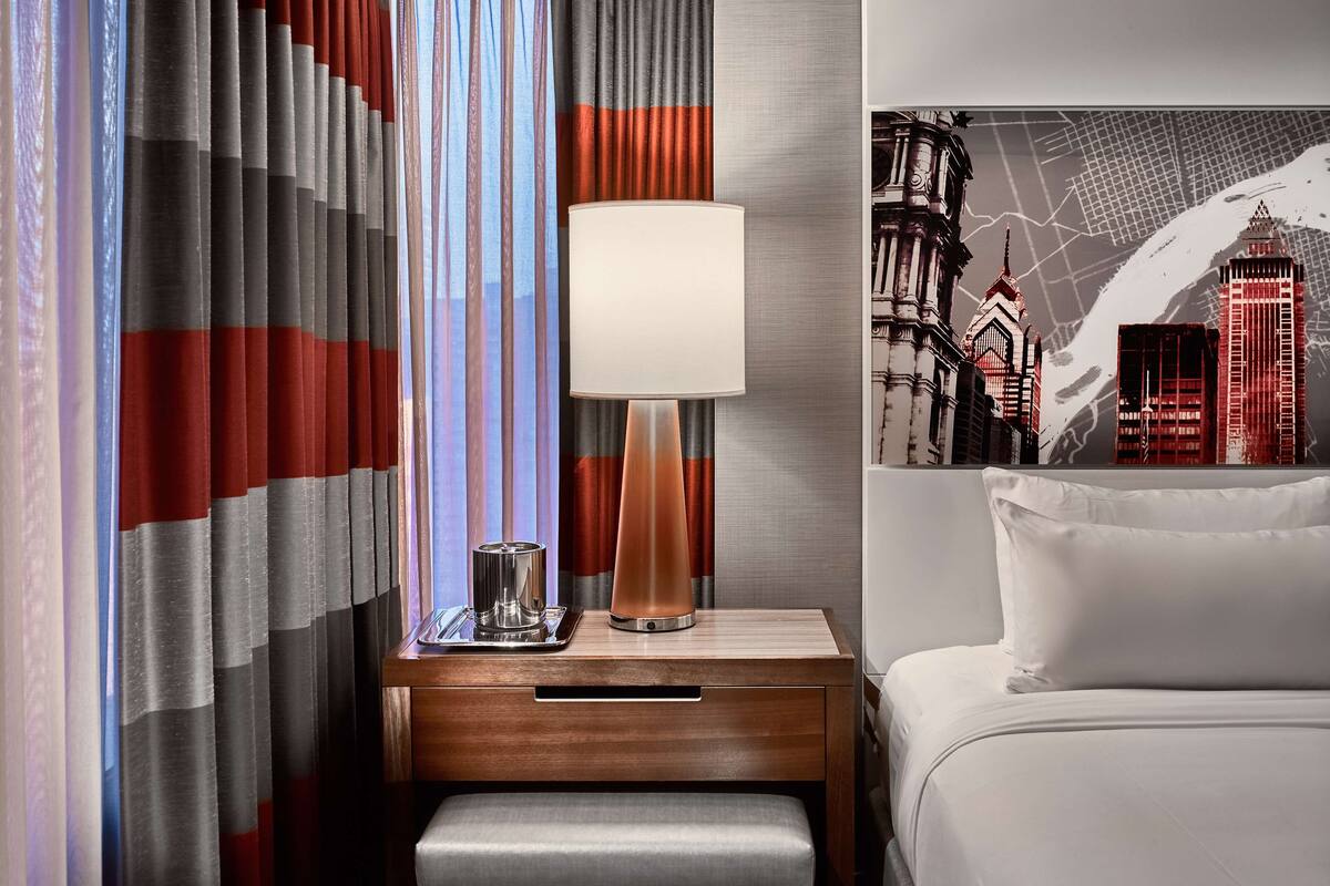 Une chambre d’hôtel avec un lit, une table de chevet avec une lampe, un banc et des rideaux.