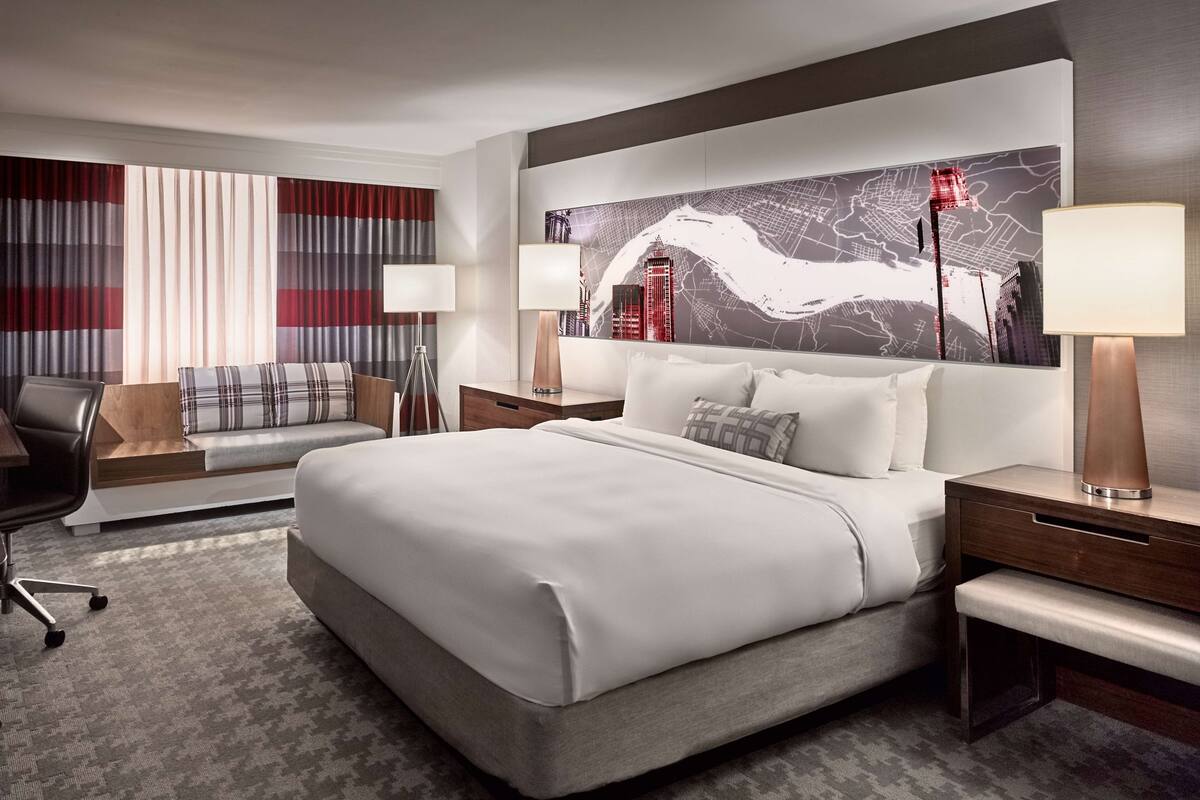 Une chambre d’hôtel moderne avec un grand lit, un canapé, un bureau et une œuvre d’art représentant un paysage urbain accrochée au mur.