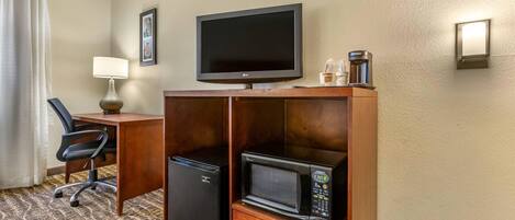 A hotel room with a wooden desk, a television, a microwave, a refrigerator, a chair, a lamp, and a coffee maker.