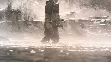 A person standing by a river, wearing a jacket and hat, with a fishing rod.