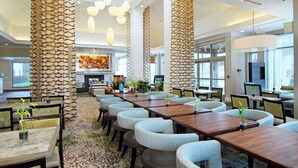 Um lobby de hotel moderno com tetos altos, colunas estampadas e uma variedade de opções de assento, incluindo mesas e cadeiras.