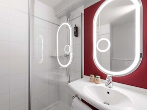 A modern bathroom with a glass-enclosed shower, a white sink with a red accent wall, and a large mirror with integrated lighting.