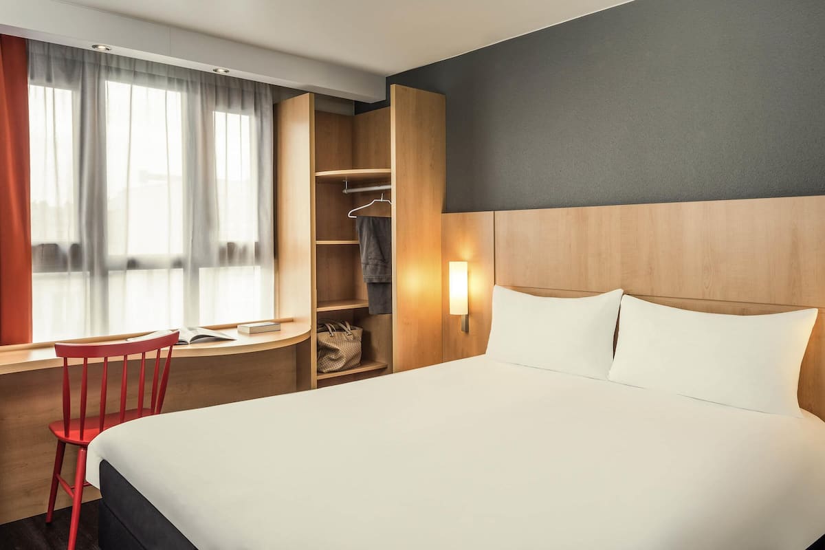 A hotel room with a bed, a desk, a chair, a wardrobe, and a window with curtains.
