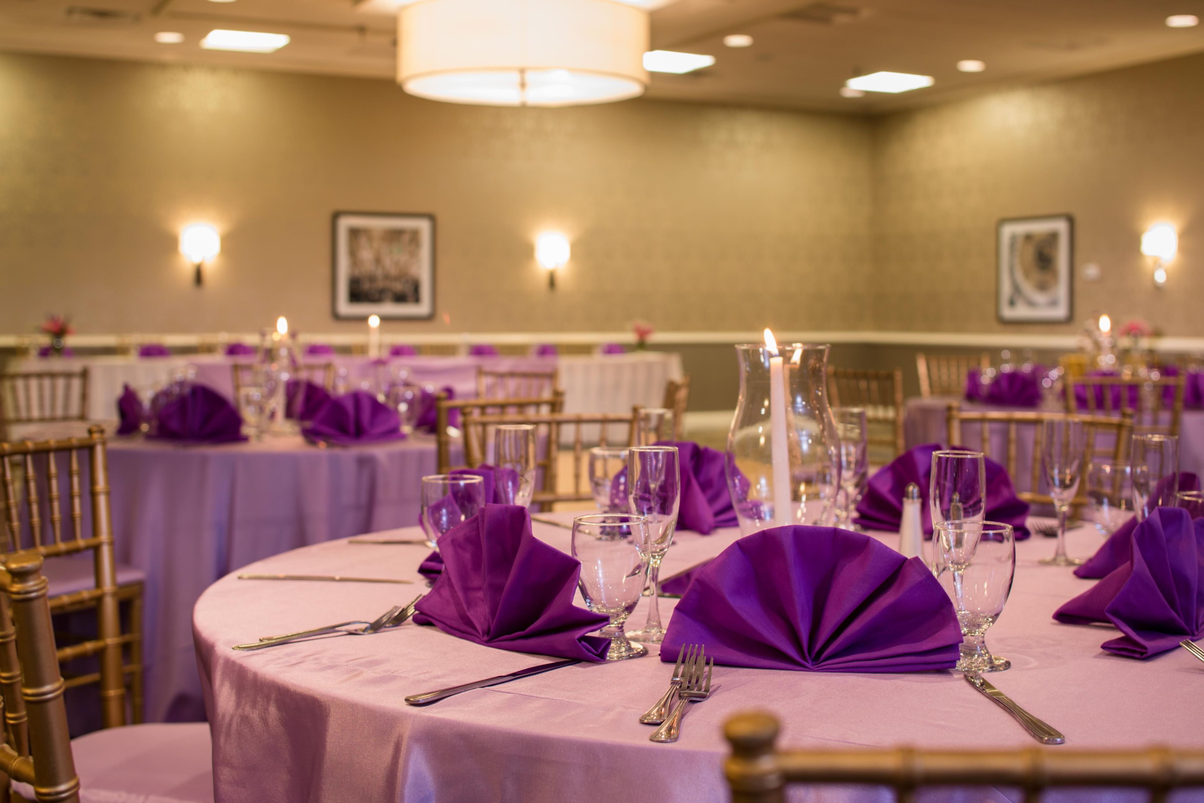 Une salle de banquet avec des tables rondes dressées pour le repas, chacune recouverte d’une nappe violette, agrémentée de couverts en argent et de verres.