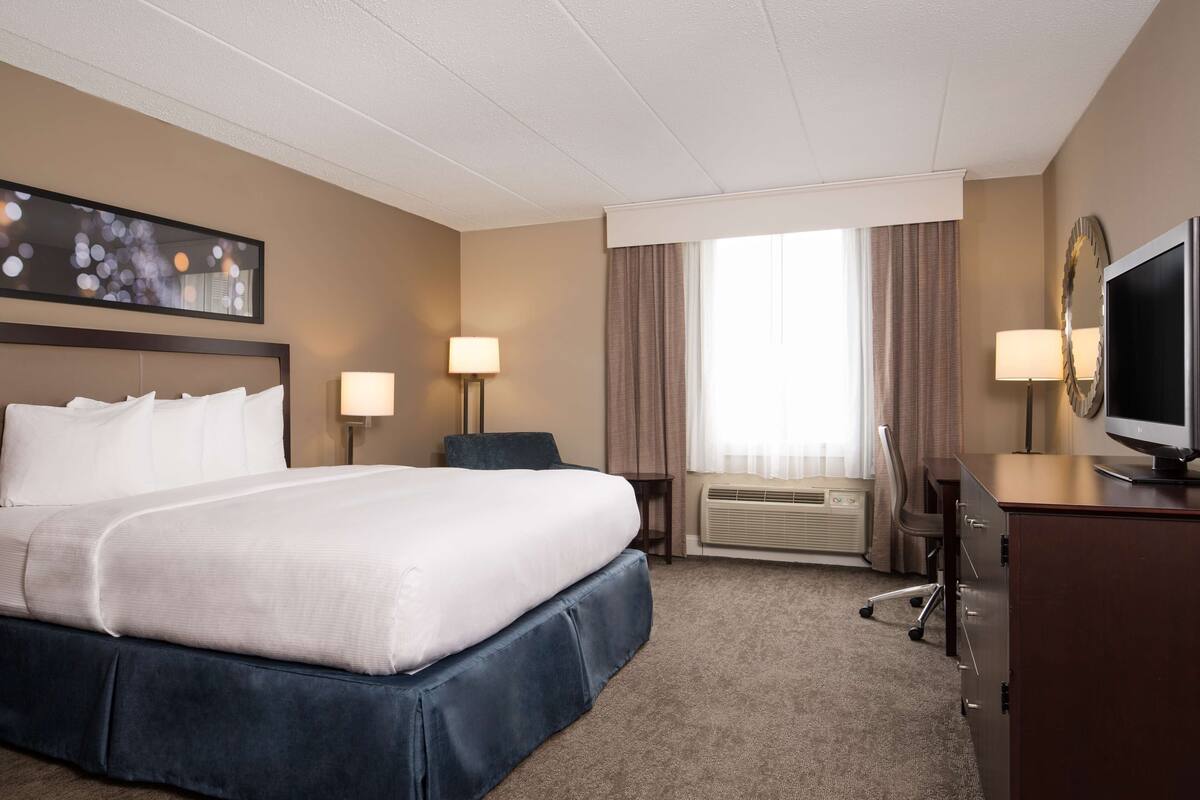 A hotel room with a large bed, two bedside tables with lamps, a desk with a chair, a television, and a large window with curtains.