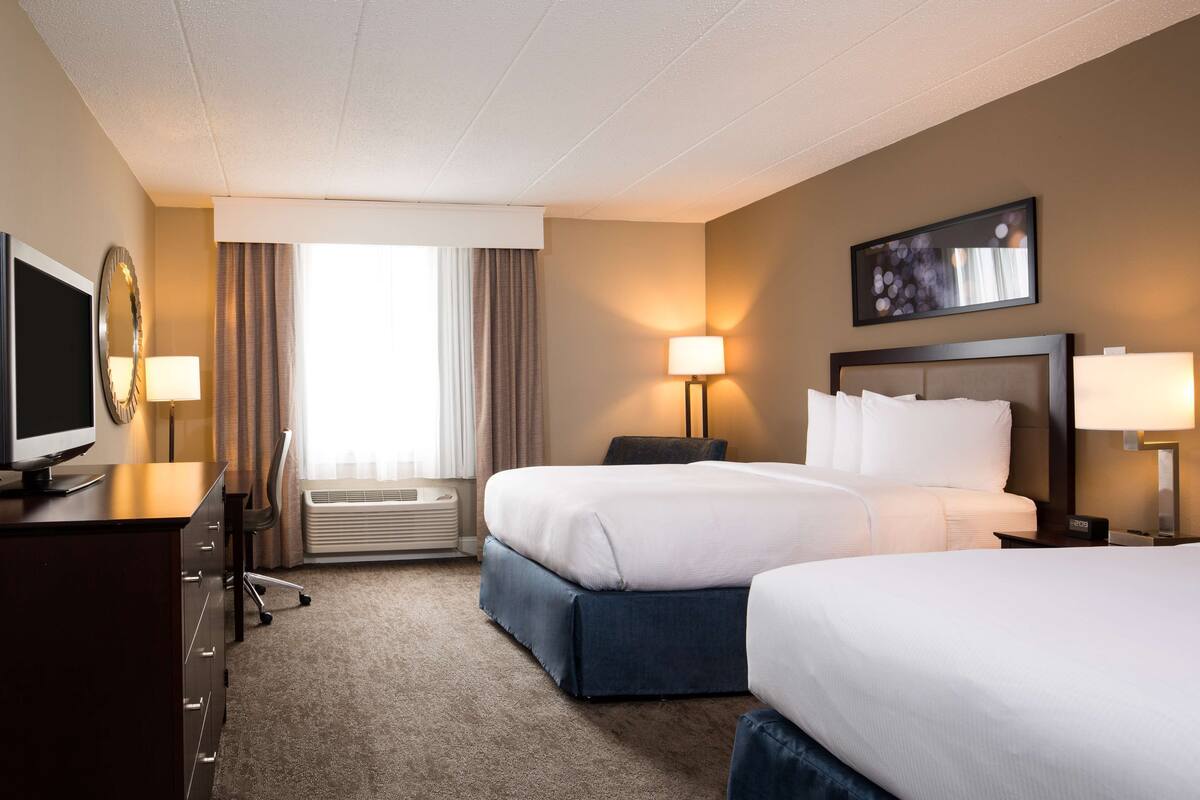 A hotel room with two beds, a desk with a TV, a chair, a lamp, and a window with curtains.
