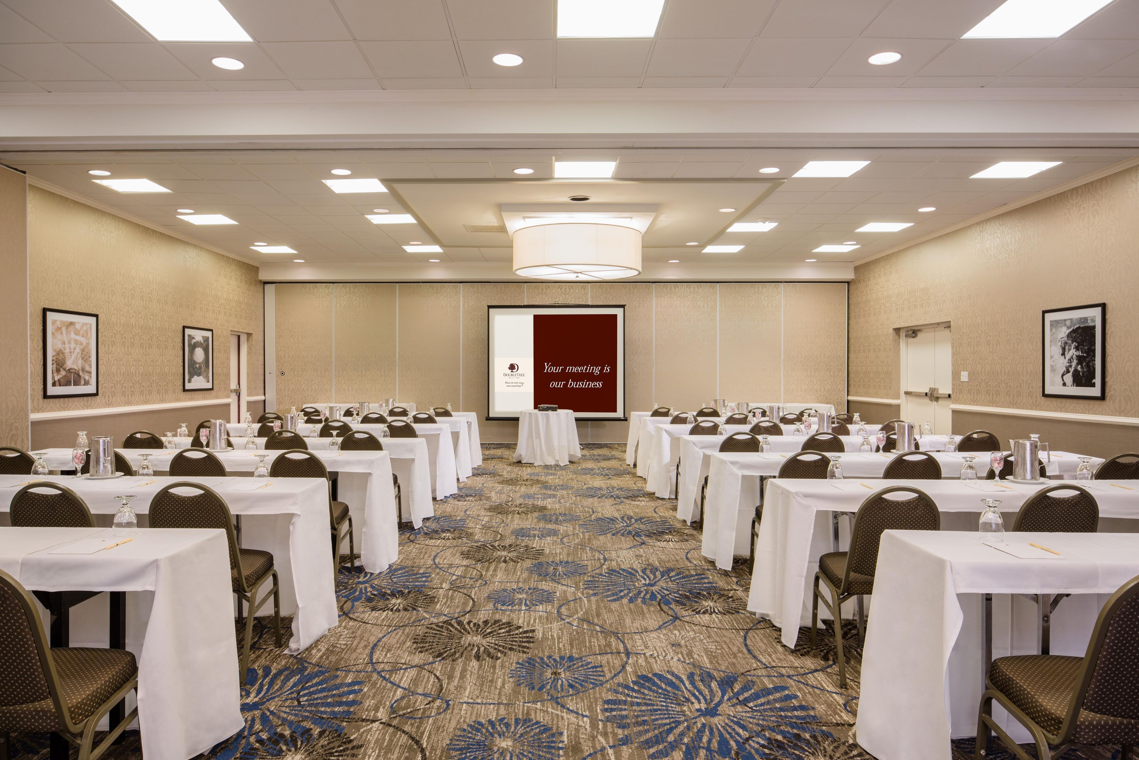 A conference room with tables and chairs, a projector screen, and a carpeted floor.