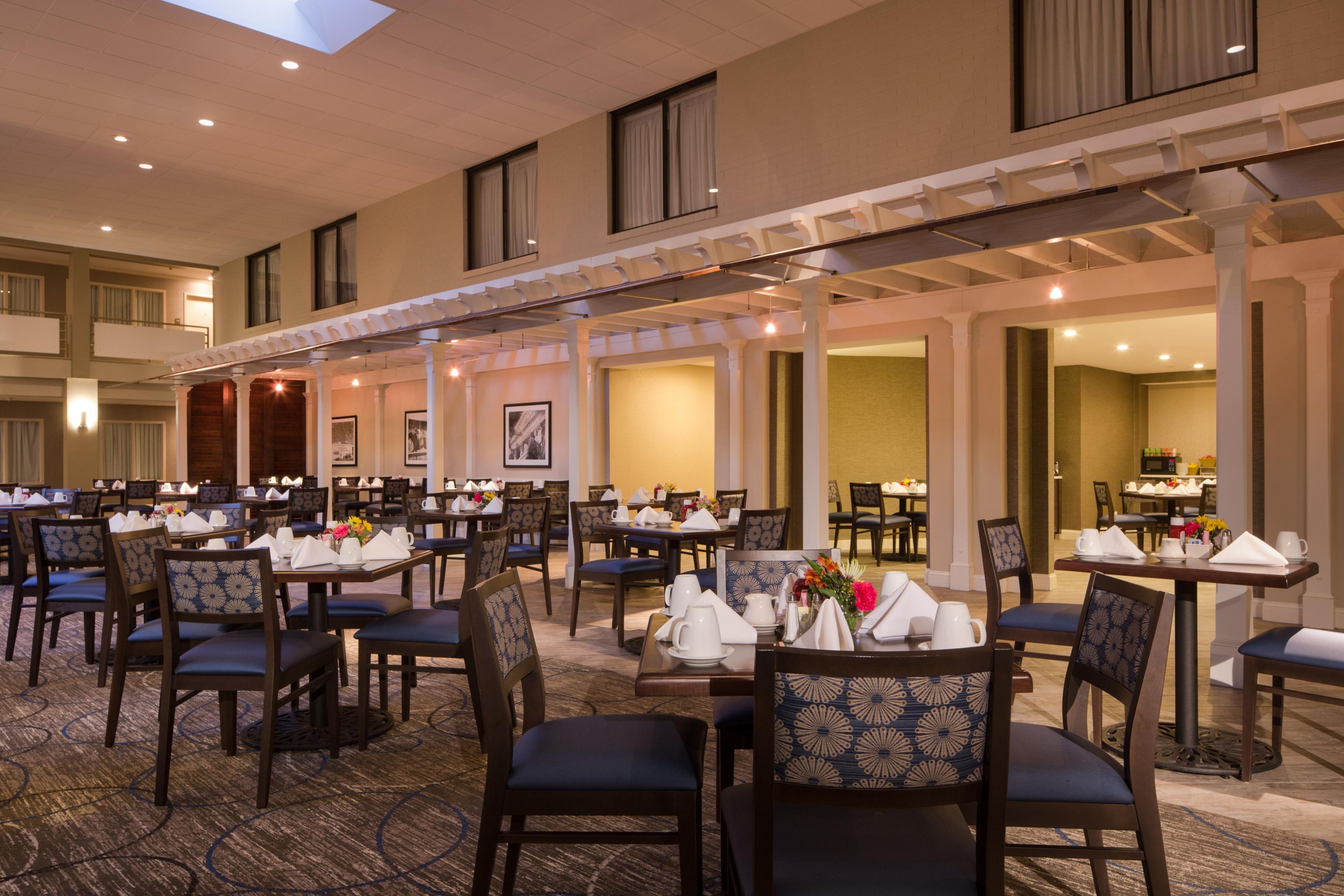 A spacious hotel dining area with multiple tables and chairs, a high ceiling with skylights, and a view of the adjacent room.