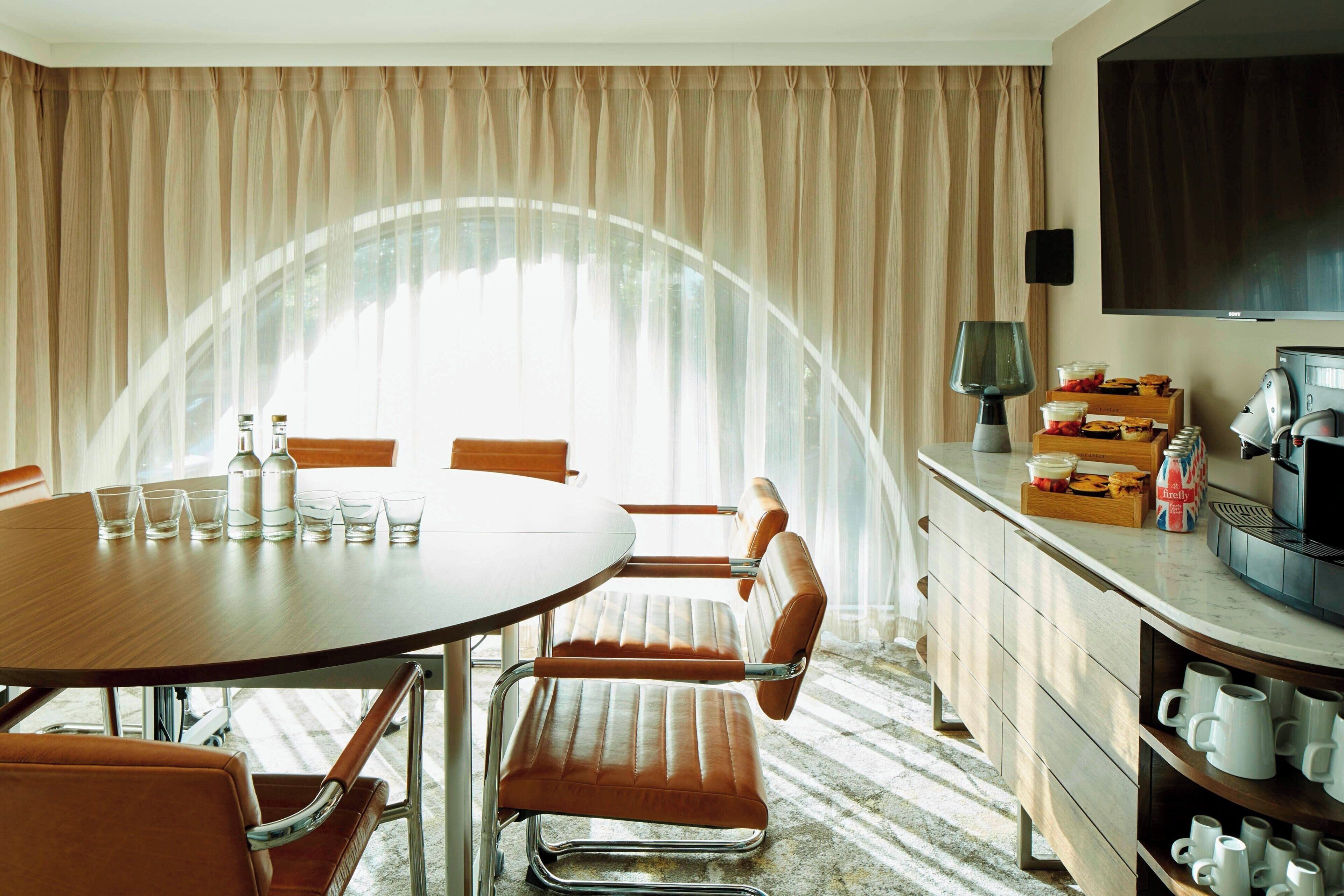 A hotel room with a round dining table, four chairs, a coffee machine, a TV, and a minibar.