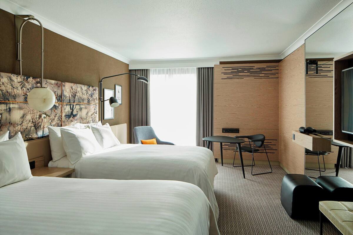 Ein Hotelzimmer mit zwei Betten, einem Schreibtisch, einem Sessel, einem Fernseher und einem großen Fenster.