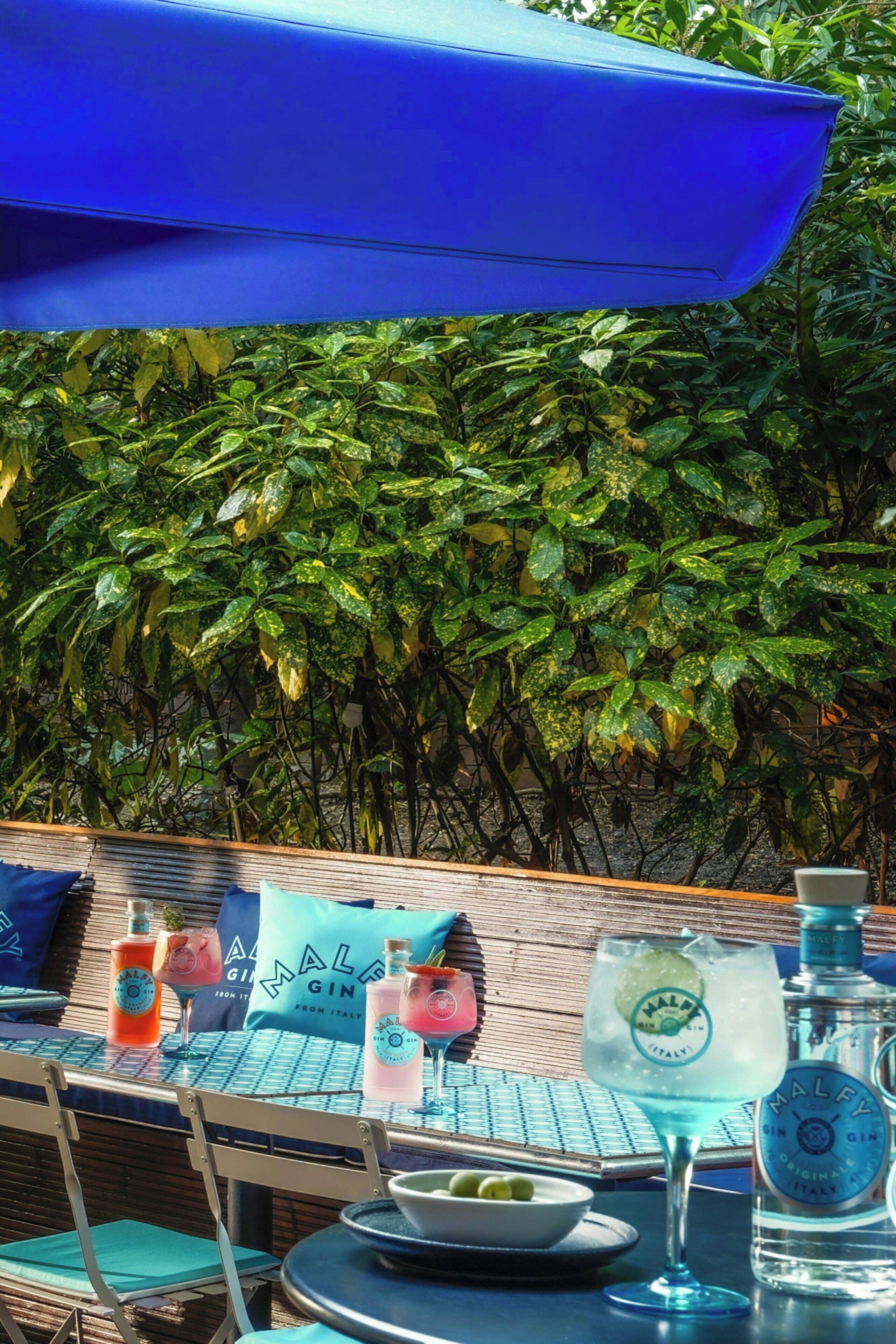 Ein Sitzbereich im Freien mit einem für zwei Personen gedeckten Tisch, inklusive eines blauen Sonnenschirms, einer Flasche Malfy Gin und einem Glas.