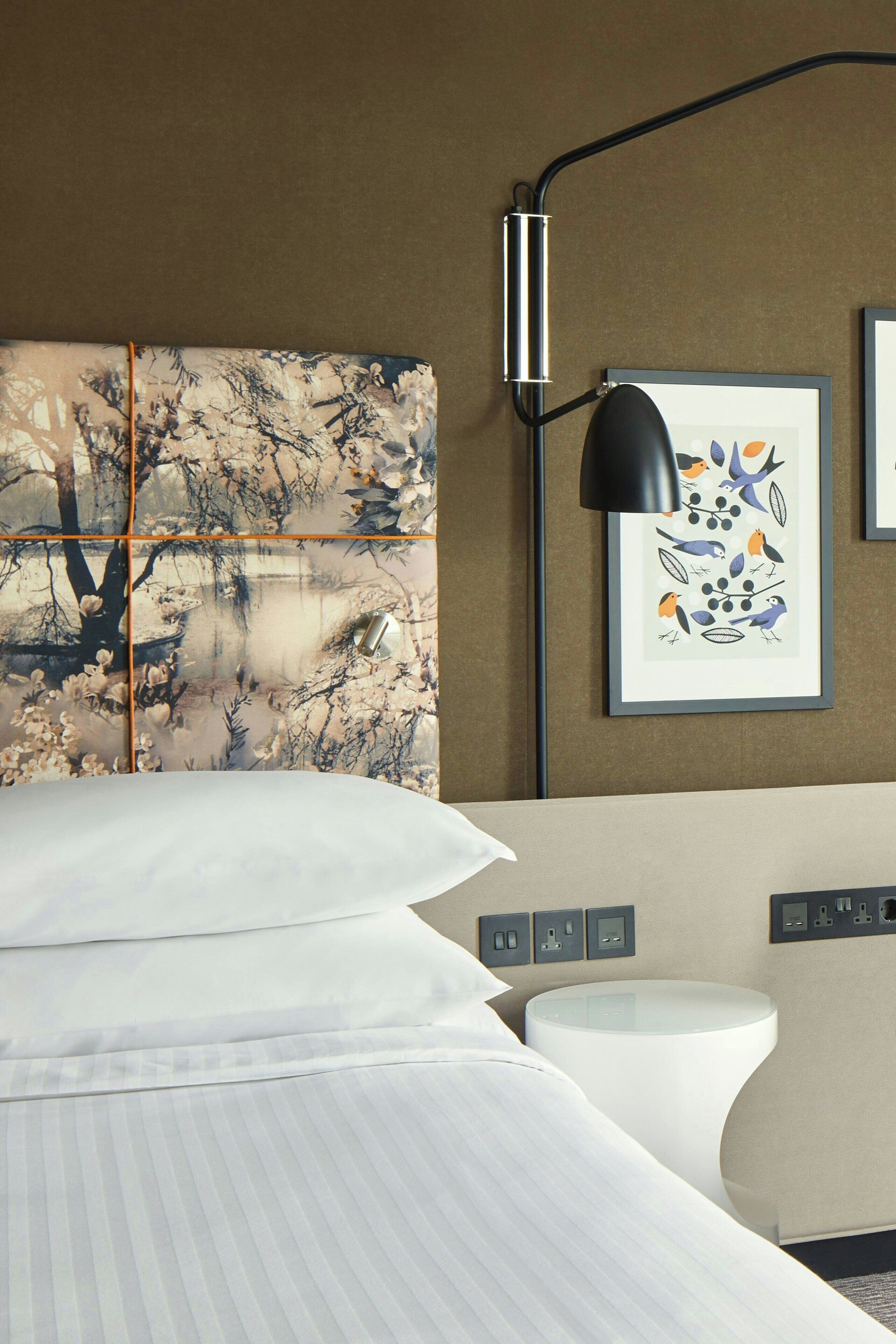 A hotel room with a large bed, a bedside table, a wall-mounted lamp, and a framed picture of birds.