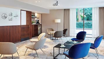 Ein modernes Hotelzimmer mit Marmorboden, einem großen Fenster mit Vorhängen, einem Projektor an der Decke und einer Sitzecke mit Stühlen und Tischen.