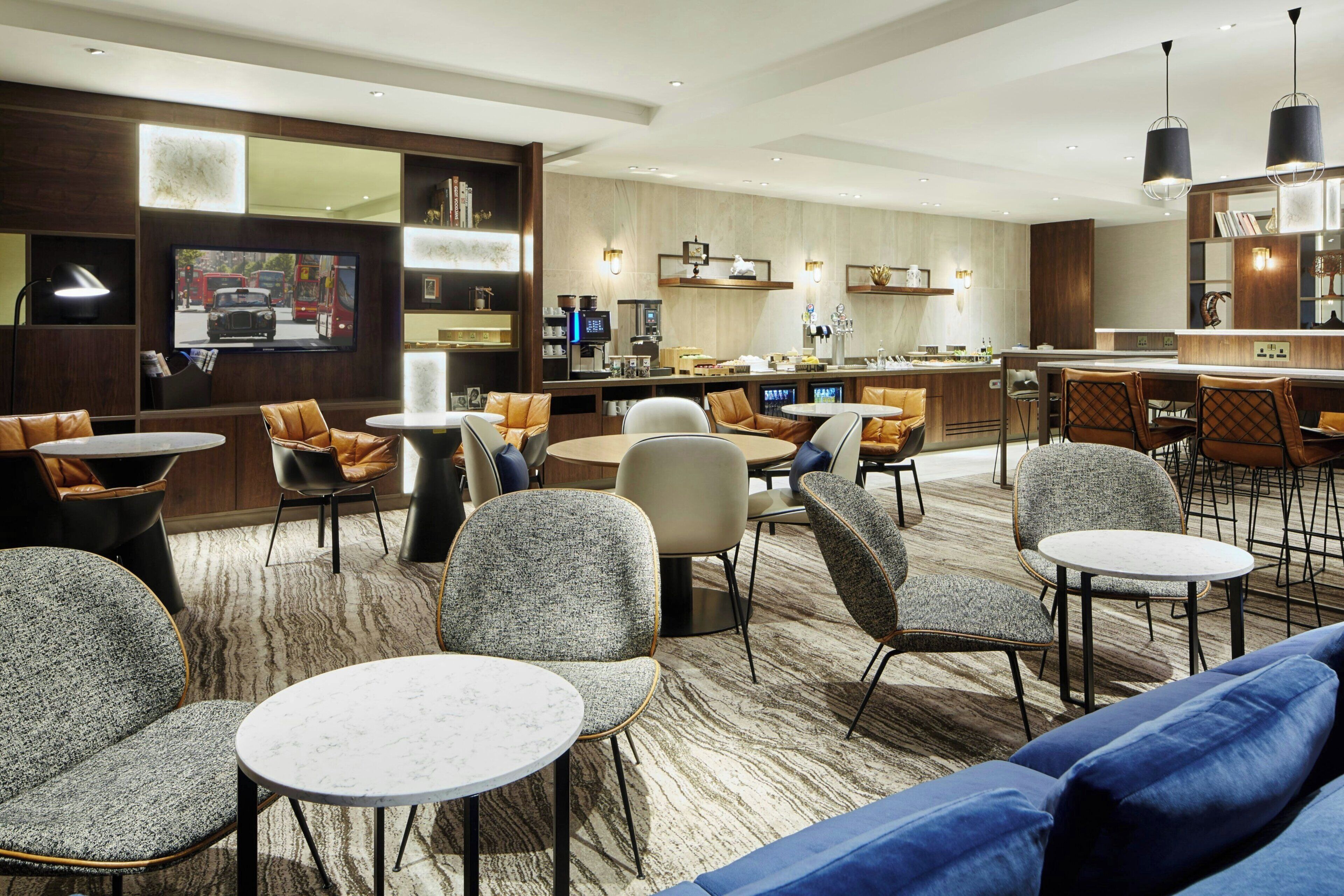 A modern hotel lobby with round tables, comfortable seating, a bar area, and a television displaying a bus.