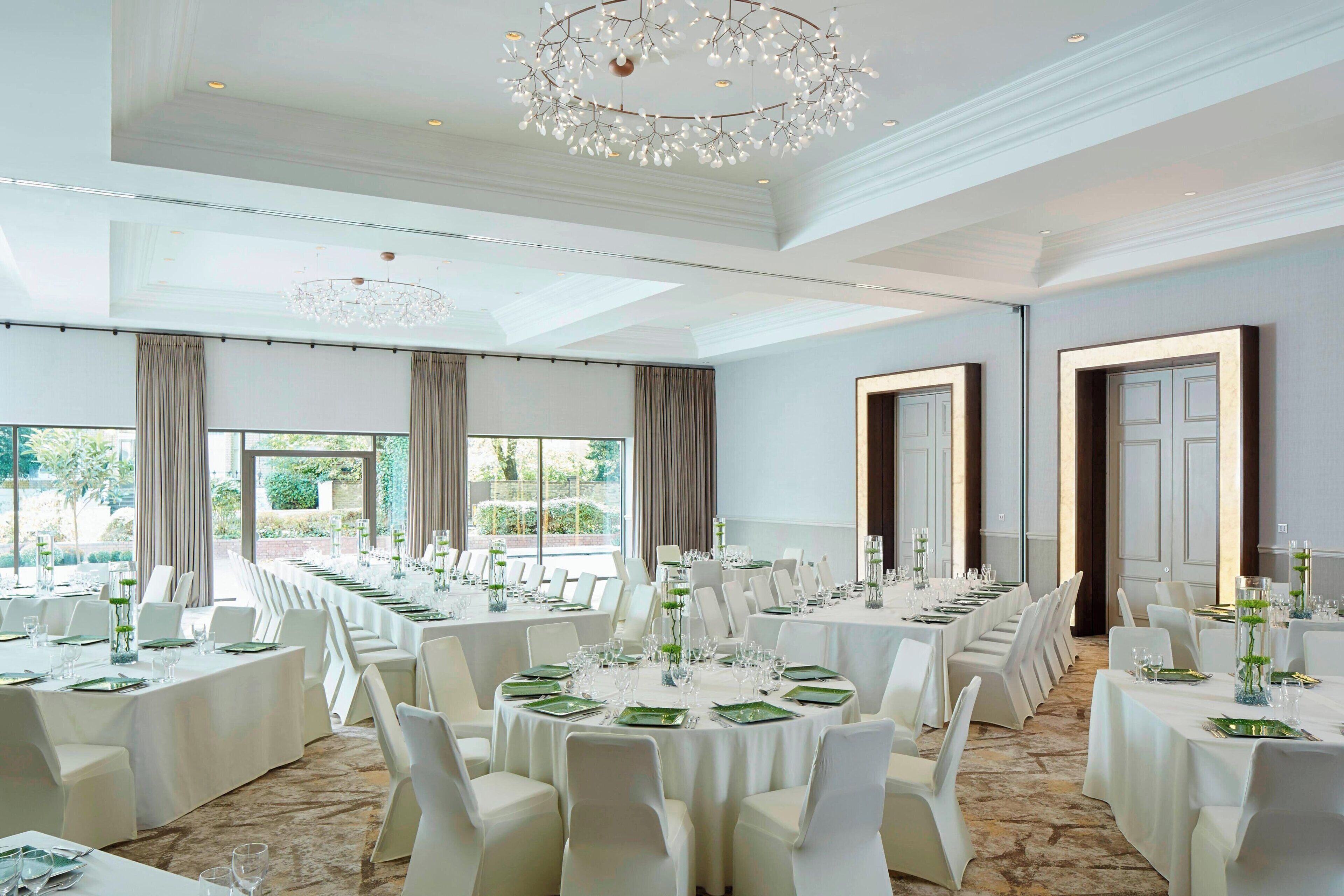 A banquet hall with multiple round tables set for dining, large windows, and a chandelier.