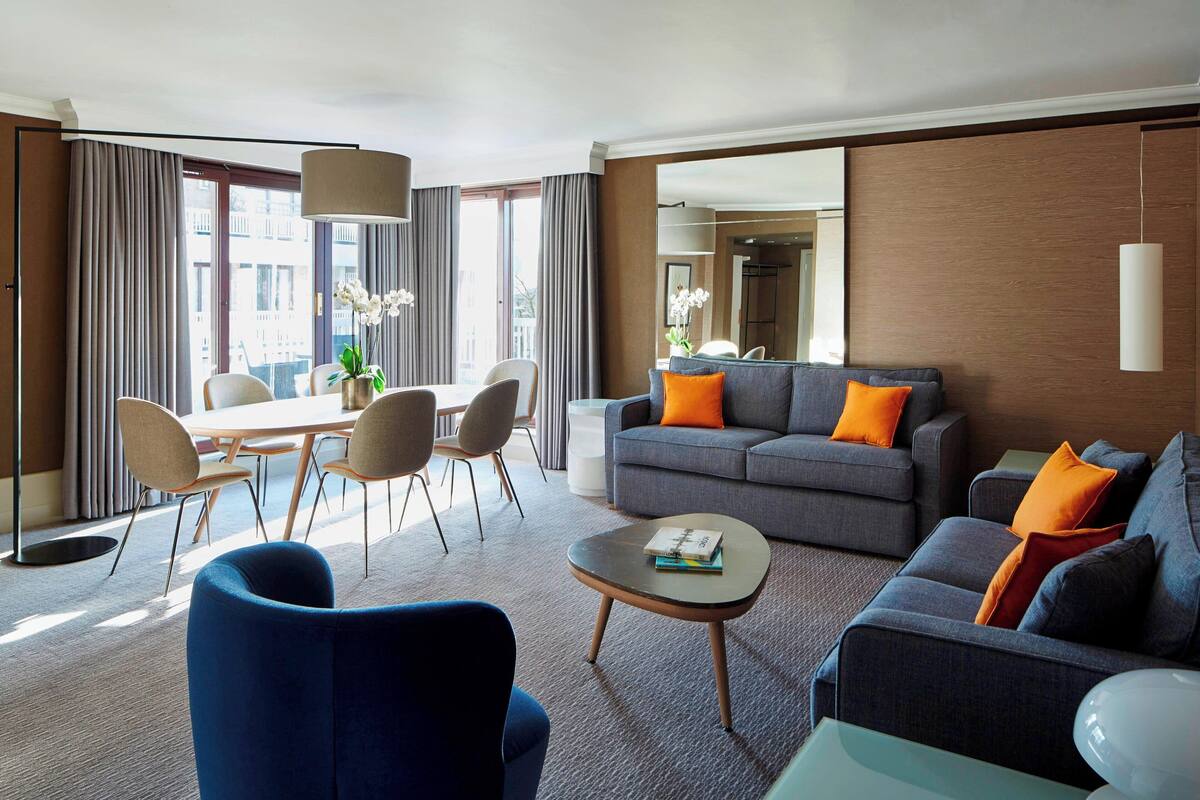 Ein modernes Hotelzimmer mit Essbereich, einer Sofagruppe und einem großen Fenster mit Vorhängen.