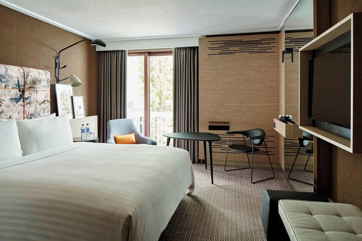 Ein Hotelzimmer mit einem großen Bett, einem Schreibtisch, zwei Stühlen, einem Flachbildfernseher und einem großen Fenster mit Vorhängen.