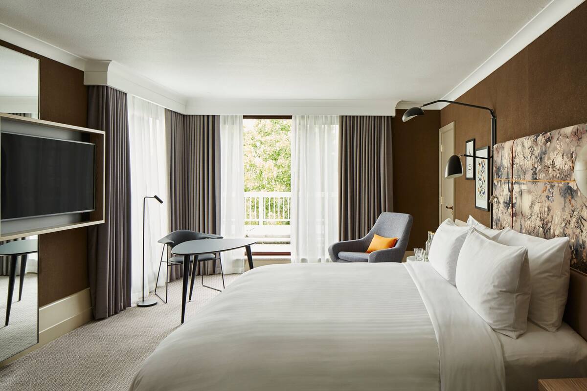 Ein Hotelzimmer mit einem großen Bett, einem Schreibtisch mit Stuhl, einem Fernseher und einem Balkon mit Vorhängen.