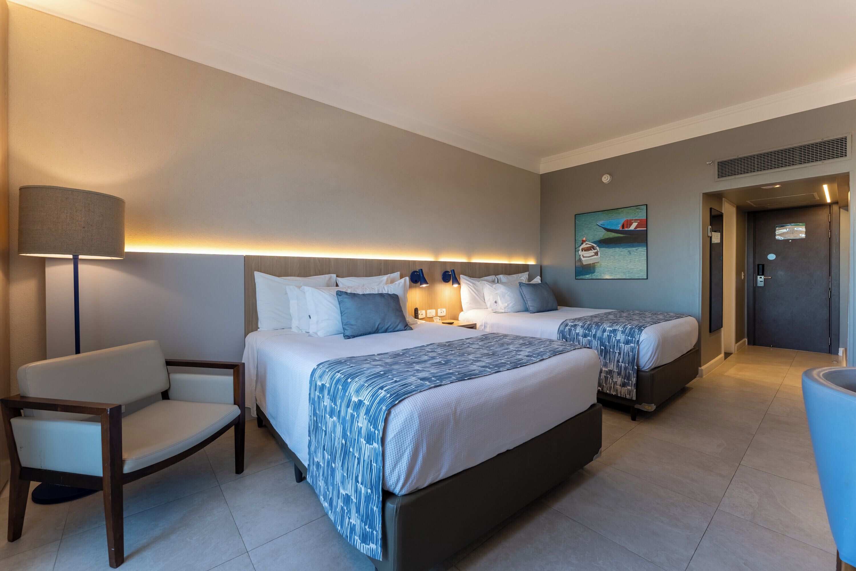 Habitación de hotel con dos camas, un escritorio, una silla, una lámpara y un cuadro en la pared.