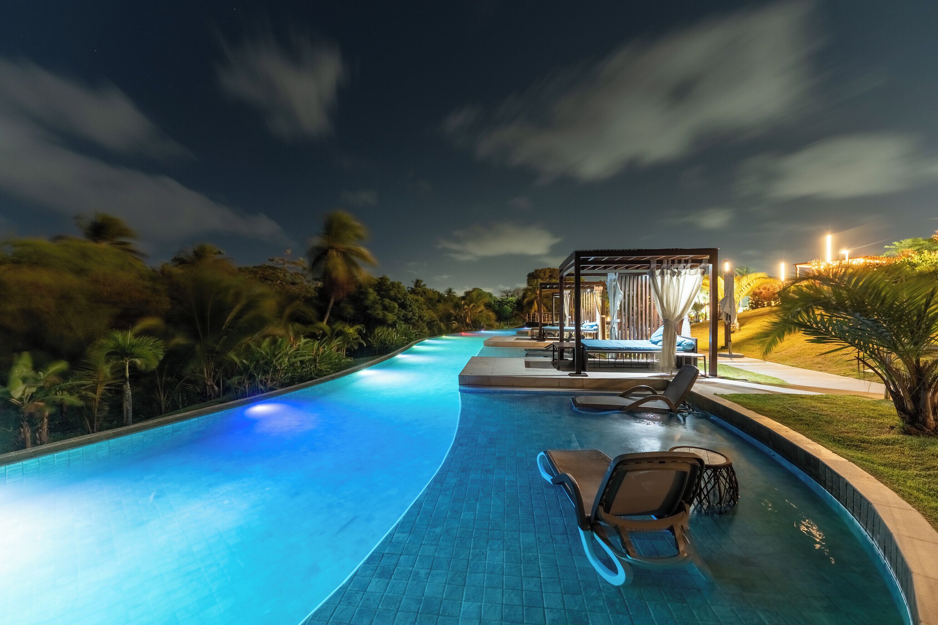 Une piscine avec des chaises longues et un espace salon couvert, éclairé le soir.