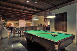 Une table de billard dans une pièce dont le plafond est orné de poutres en bois, avec un coin bar et une grande plante en pot.
