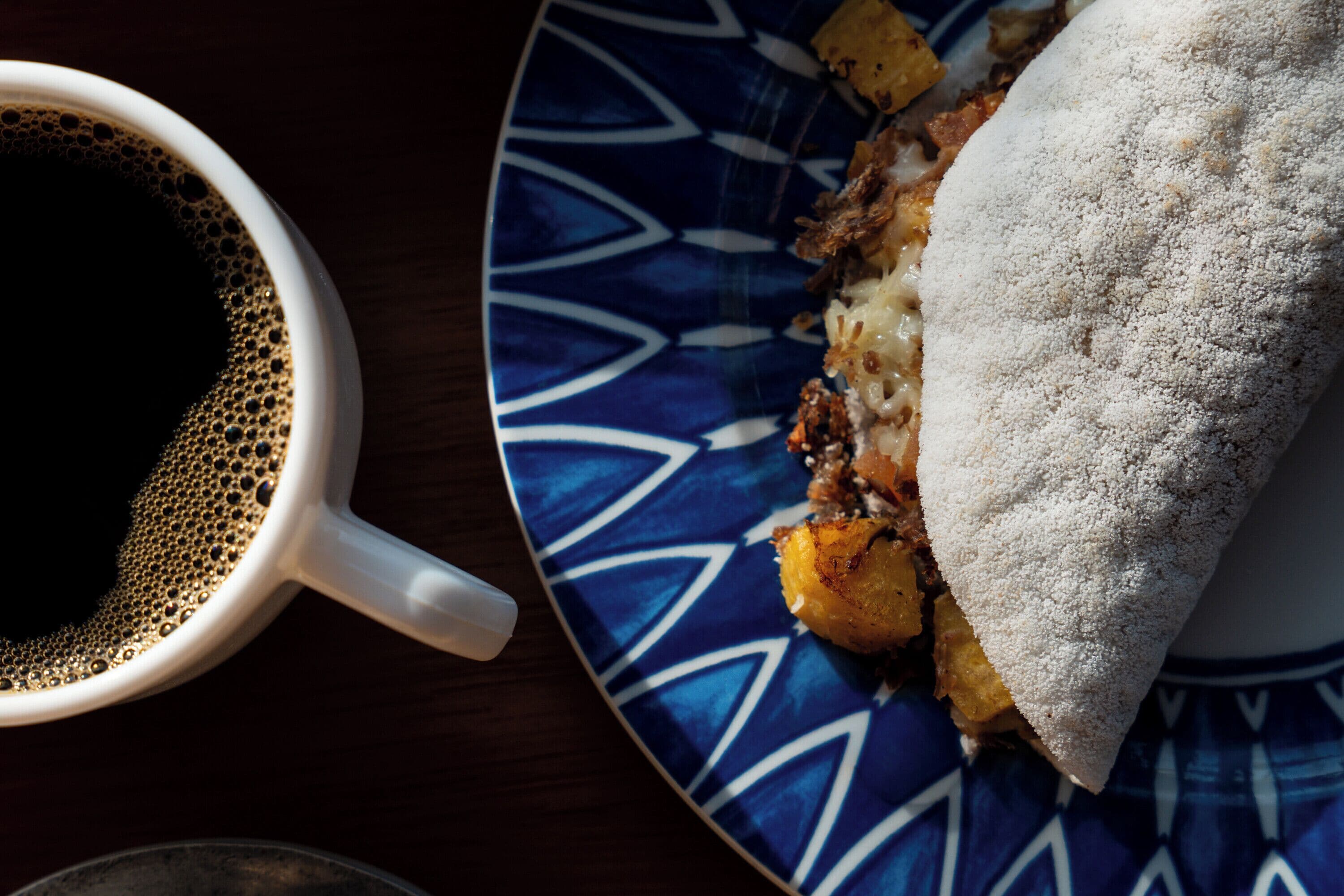 A cup of coffee and a plate with food, including a sandwich and potatoes.