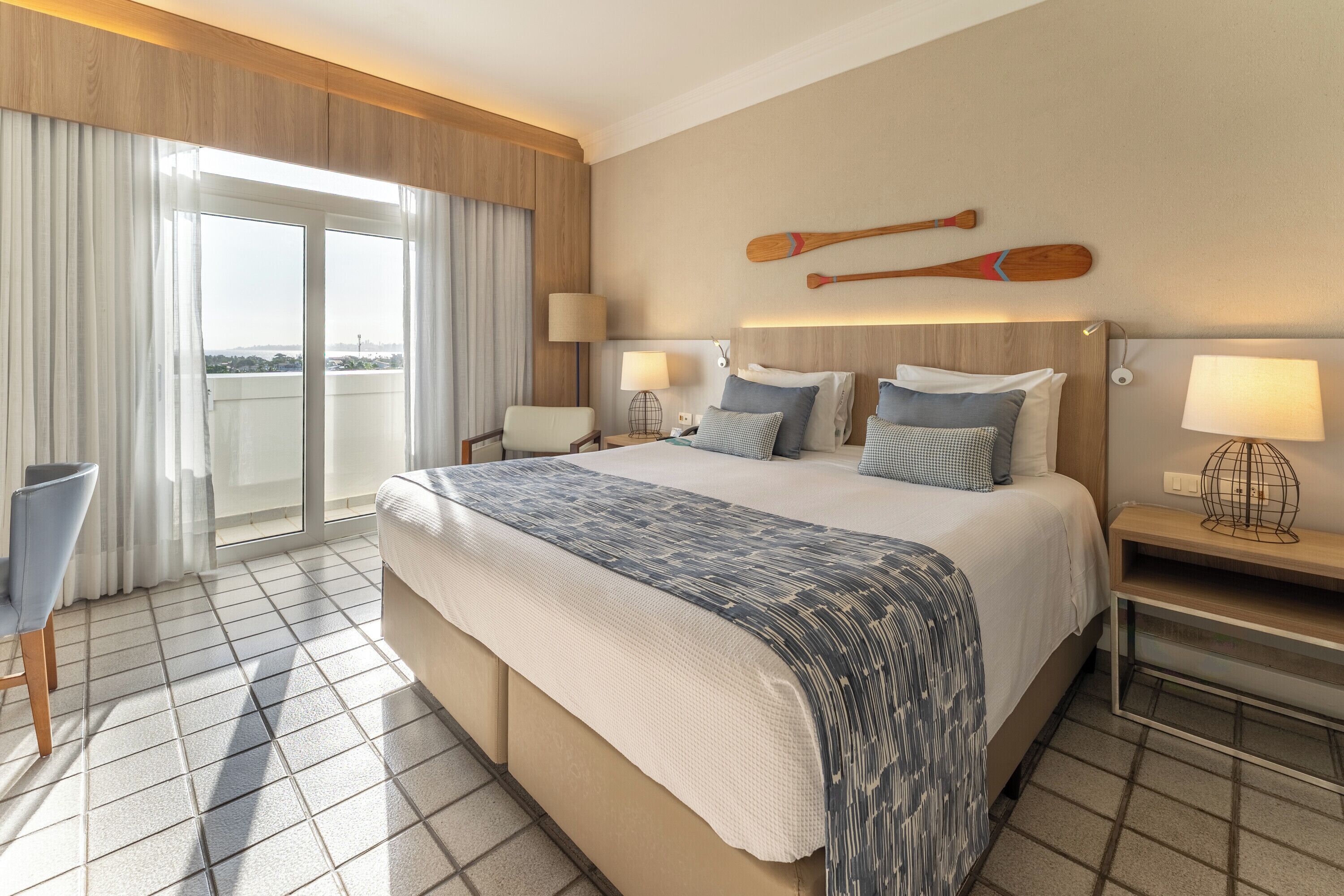 Habitación de hotel con una cama grande, dos mesitas de noche, una silla, vista a la ciudad y un balcón con cortinas.