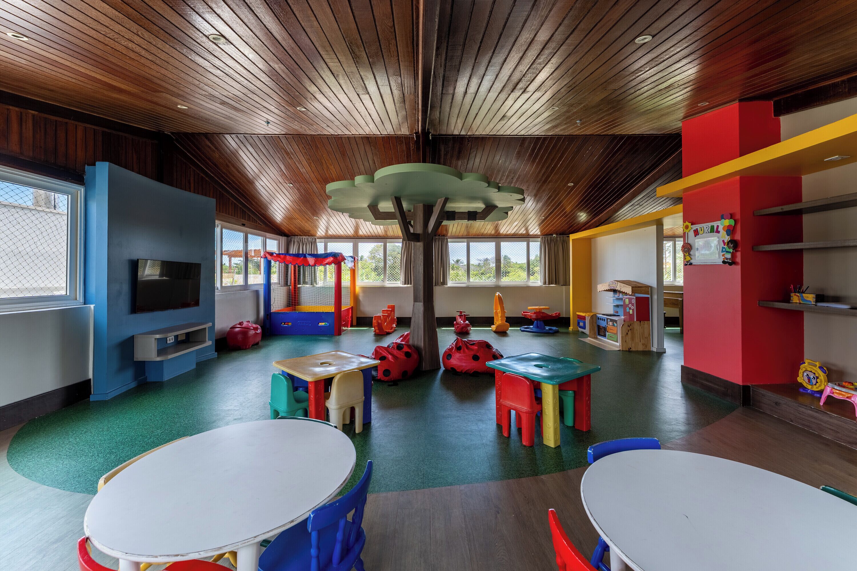 A children's playroom with wooden ceiling, colorful play tables, and a TV.