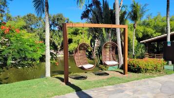 Um conjunto de baloiço de madeira com duas cadeiras de vime num jardim exuberante com um lago e plantas tropicais.