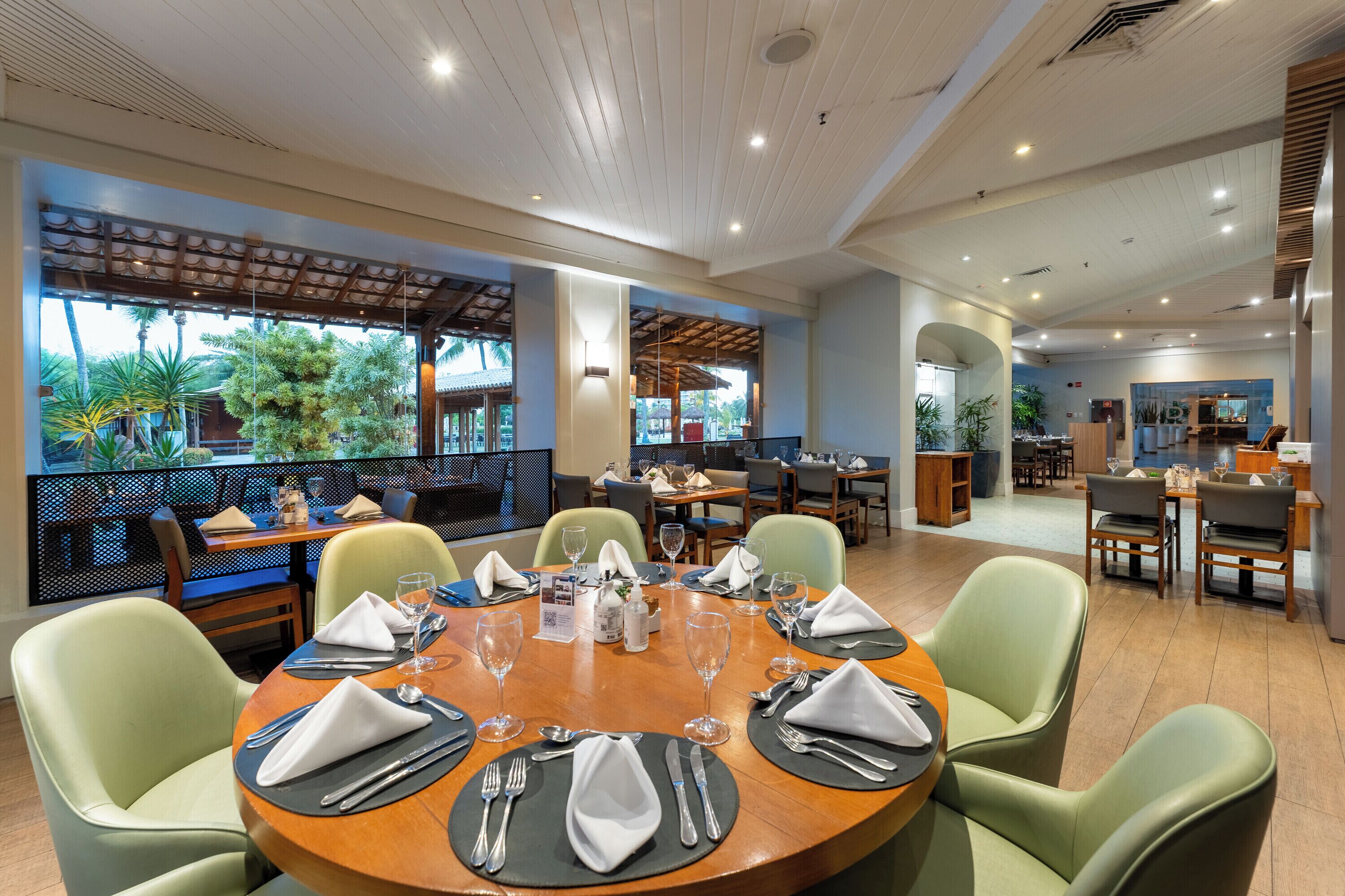 A restaurant with round tables, green chairs, and a view of palm trees through large windows.